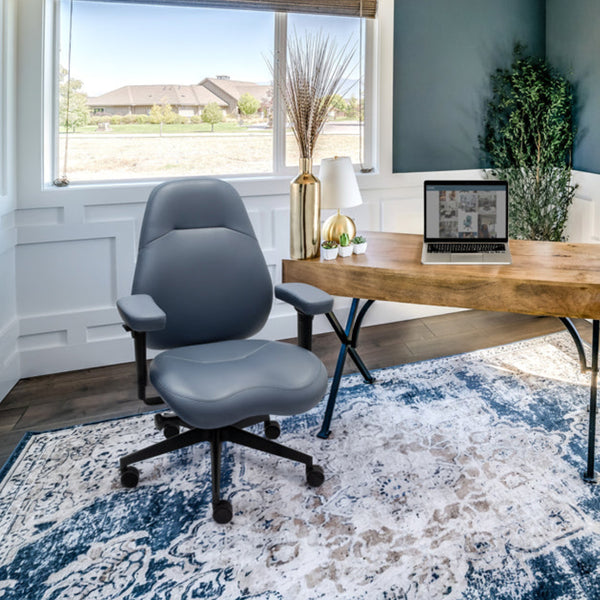 A modern home office interior with a wooden writing desk, gray office chair, patterned blue and white area rug, dark slate blue walls, white wainscoting, and large windows with brown woven shades.